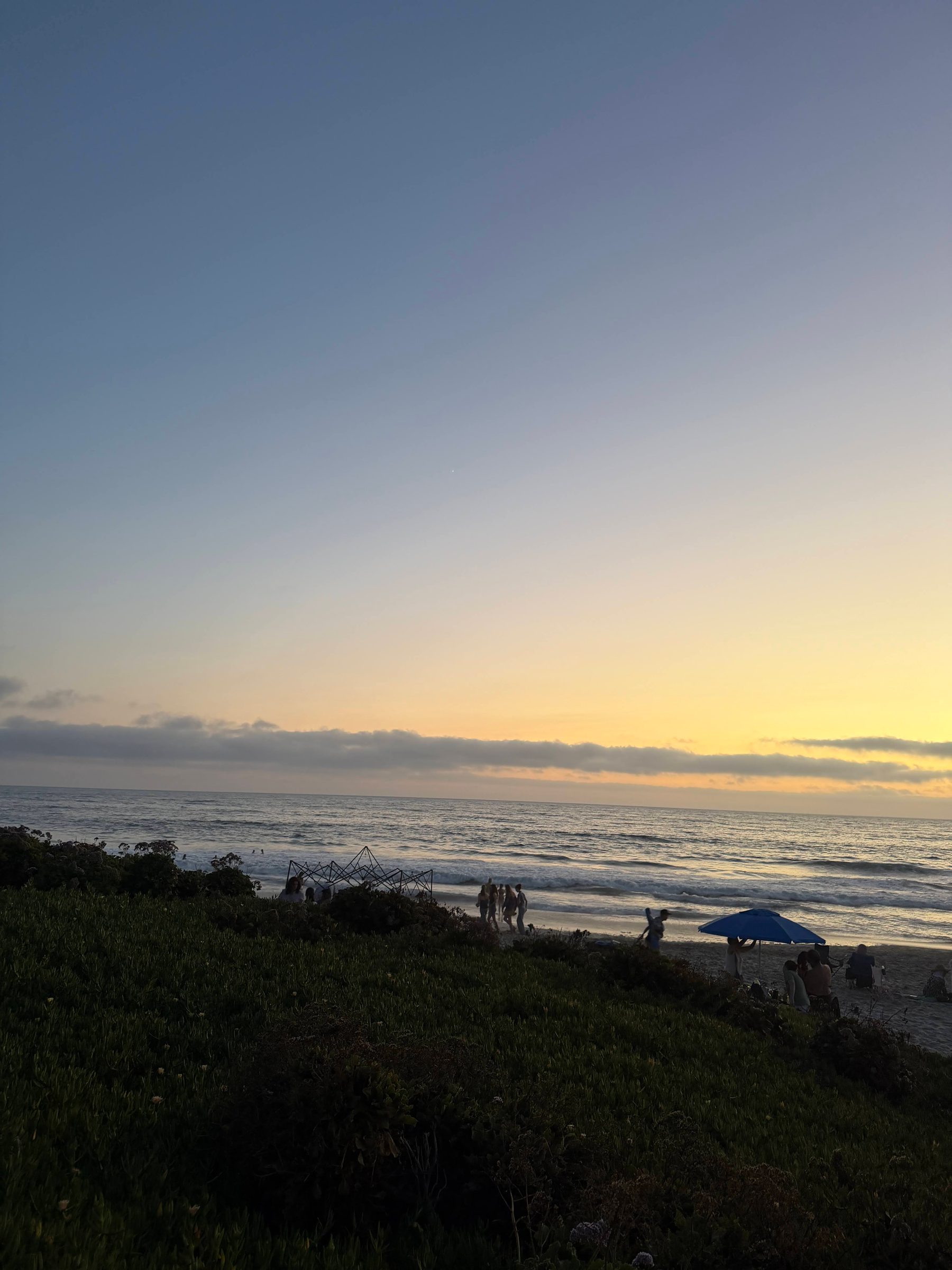A San Diego beach at sunset, people at the water's edge and a blue umbrella on the sand, the sky fading from gold to blue.